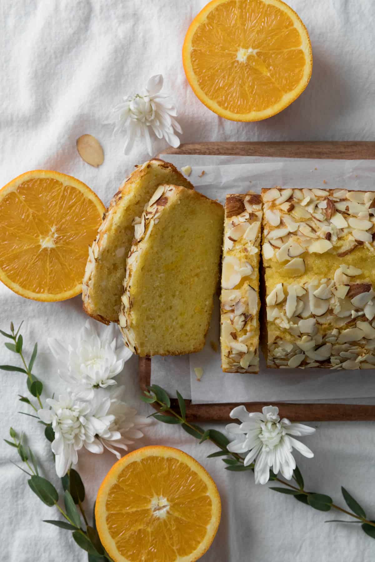 Orange Almond Loaf Cake with 3 slices cut on a wooden board with parchment paper with 3 cut oranges, white flowers, and greenery on a napkin white surface.
