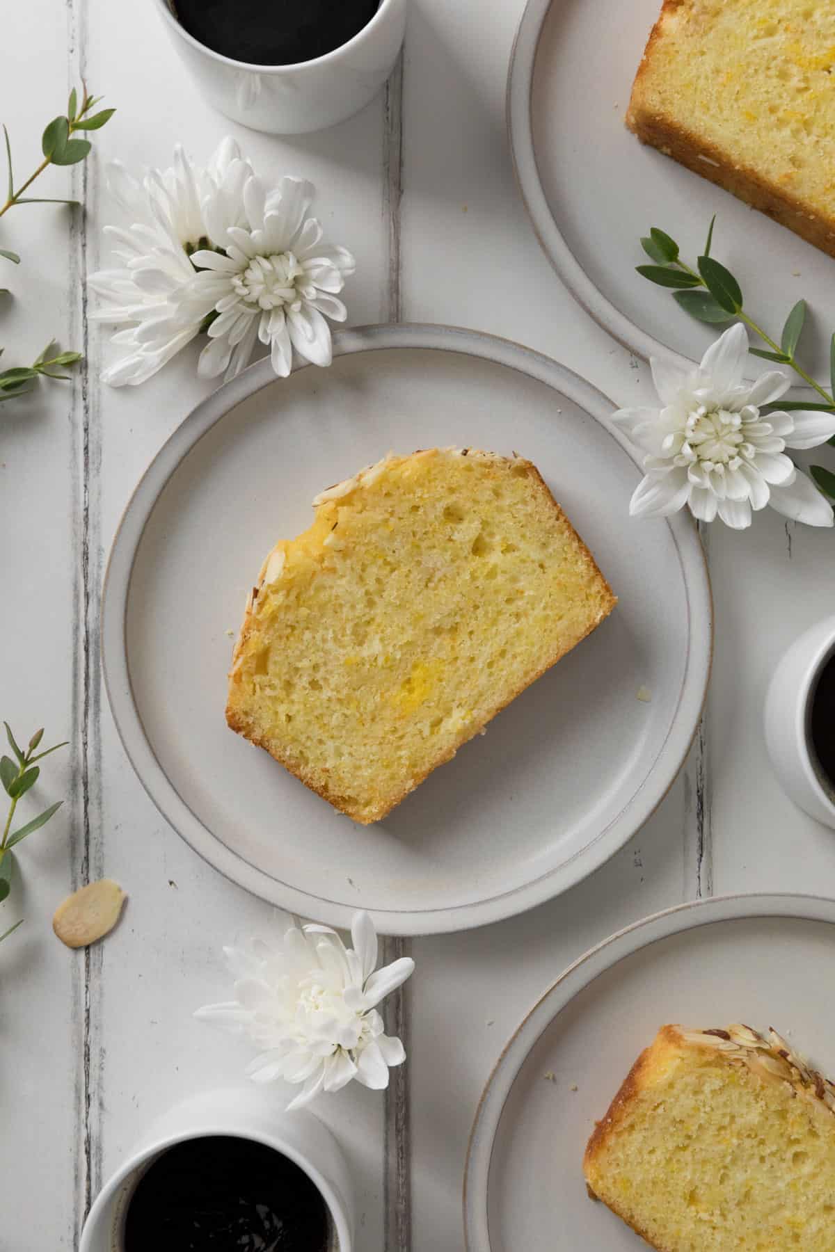 Three slices of Orange Almond Loaf Cake on individual plates surrounded by flowers, greenery, and three white espresso mugs filled with coffee on a white wood surface.