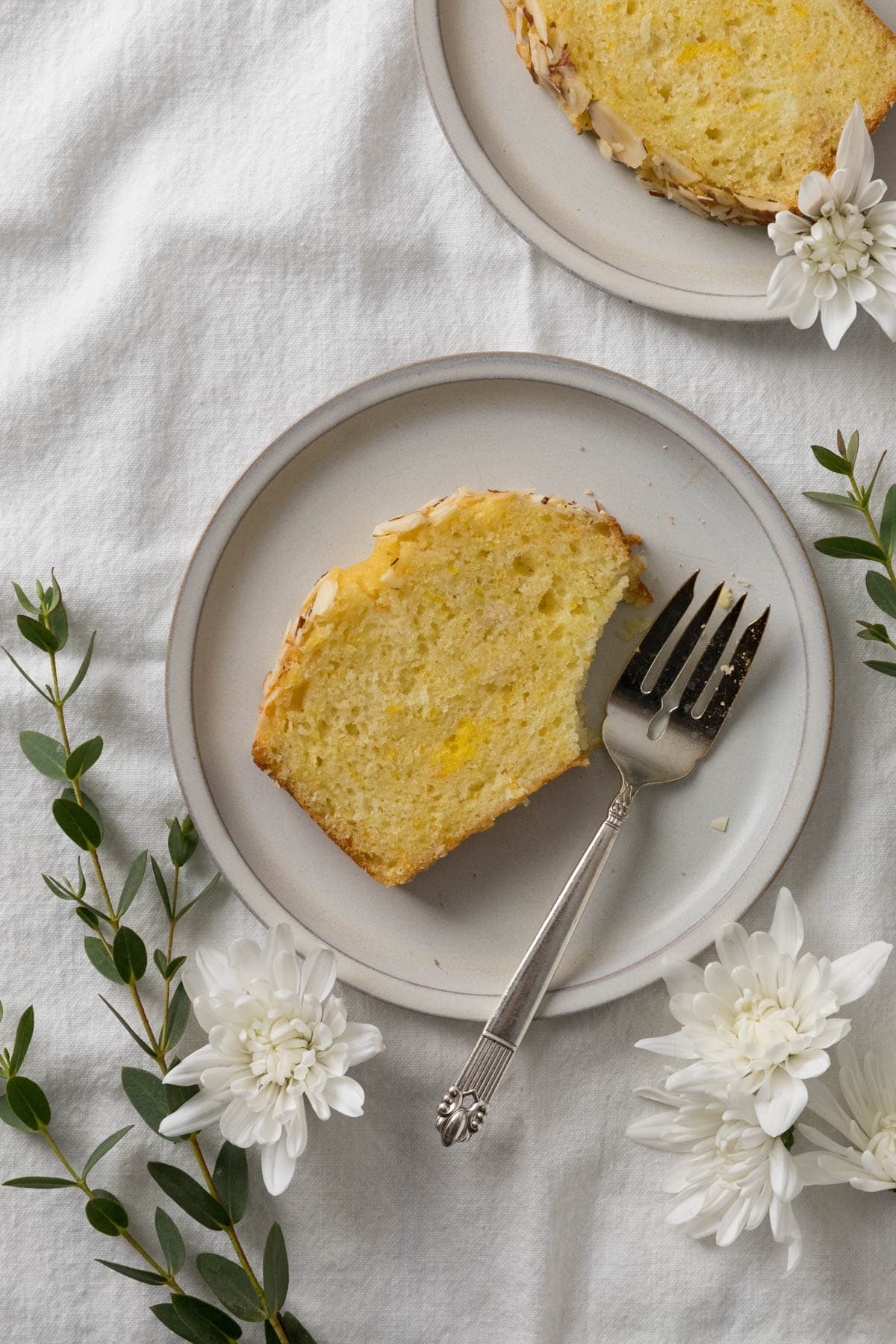 Two slices of Orange Almond Loaf Cake on individual plates with a bite taken out of one with a silver fork and surrounded by flowers and greenery on a napkin white surface.