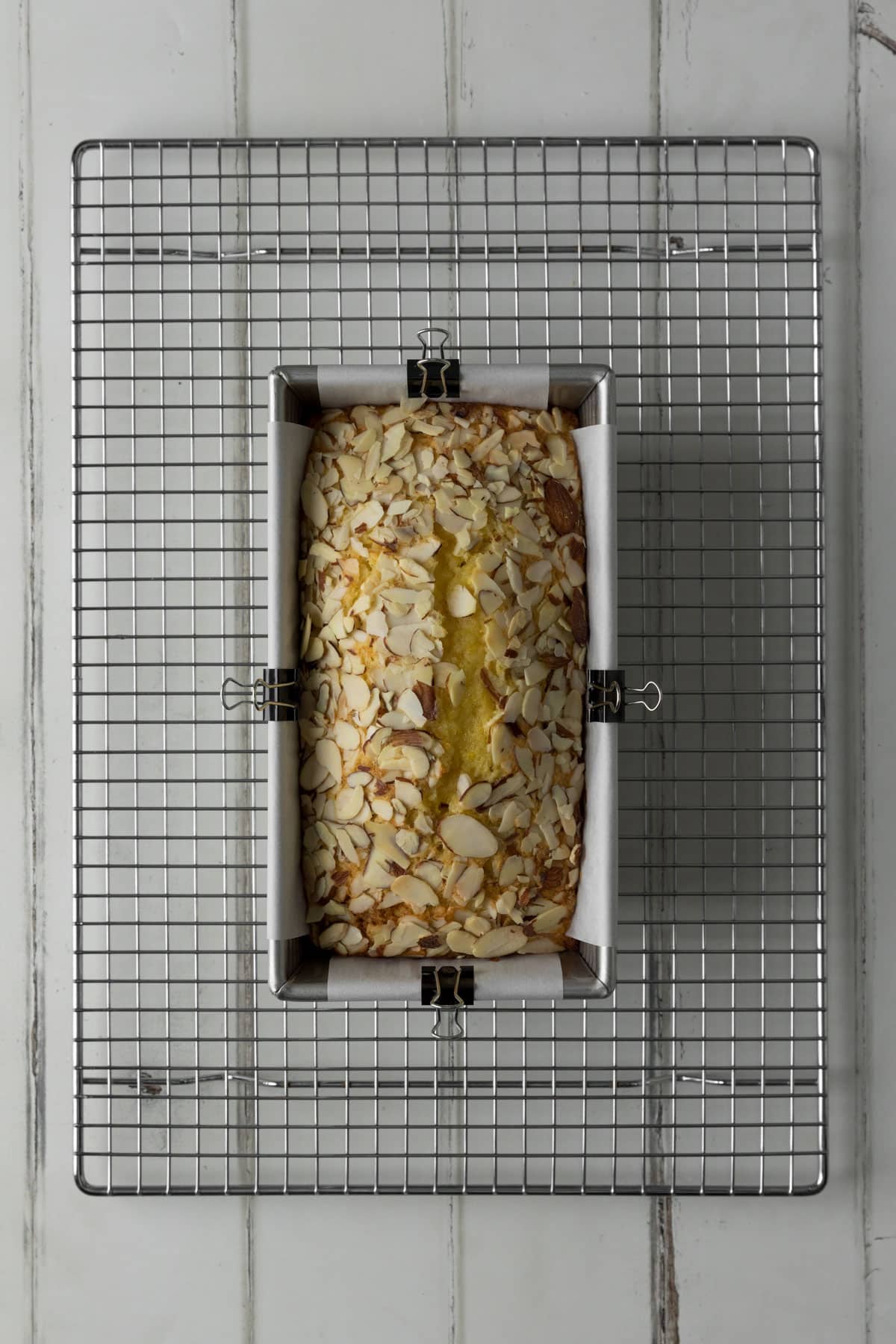 Baked Orange Almond Loaf Cake in a silver loaf pan lined with white parchment paper, secured with mini binder clips, and placed atop a silver wire cooling rack on a white wood surface.