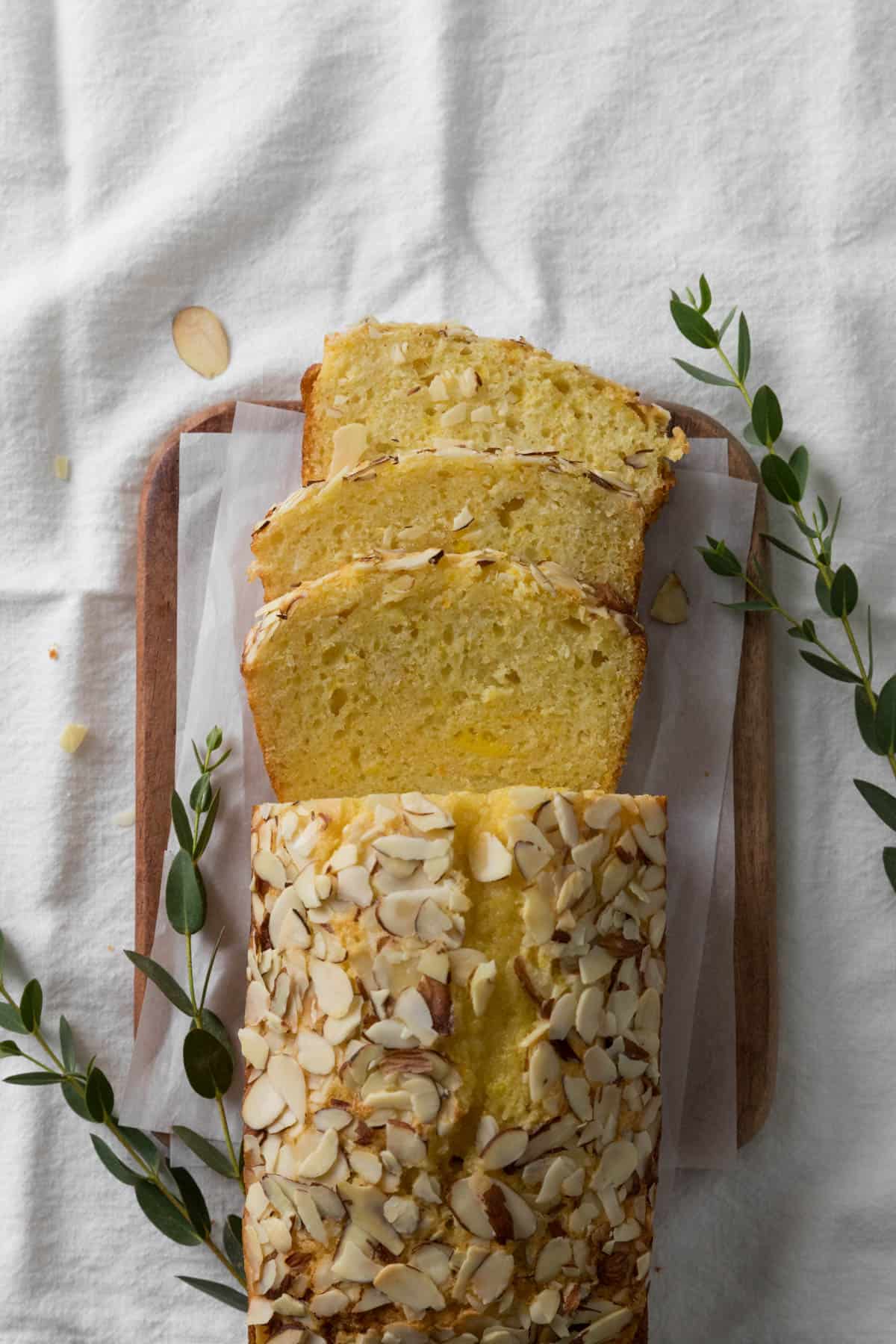 Orange Almond Loaf Cake with 3 slices cut on a wooden board with parchment paper with greenery on a napkin white surface.