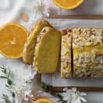 Orange Almond Loaf Cake with 3 slices cut on a wooden board with parchment paper with 3 cut oranges, white flowers, and greenery on a napkin white surface.