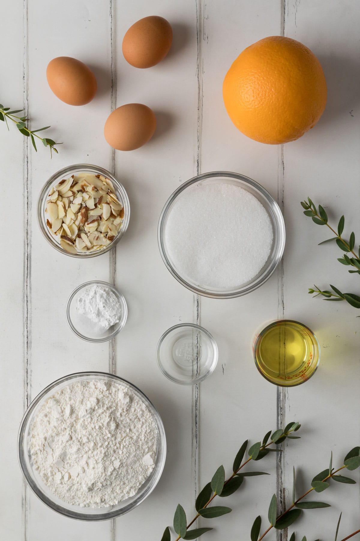 Ingredients needed to make Orange Almond Loaf Cake displayed in clear bowls on a white wood surface with greenery.