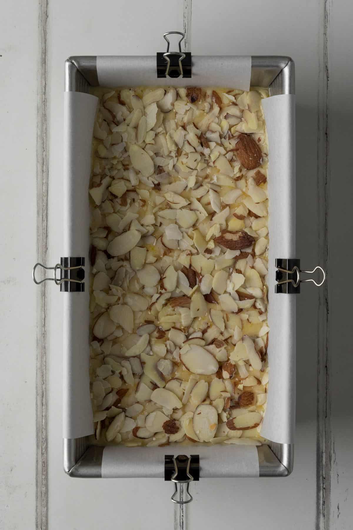 Silver loaf pan lined with white parchment paper, secured with mini binder clips, filled with orange almond loaf cake batter, and topped with sliced almonds on a white wood surface.