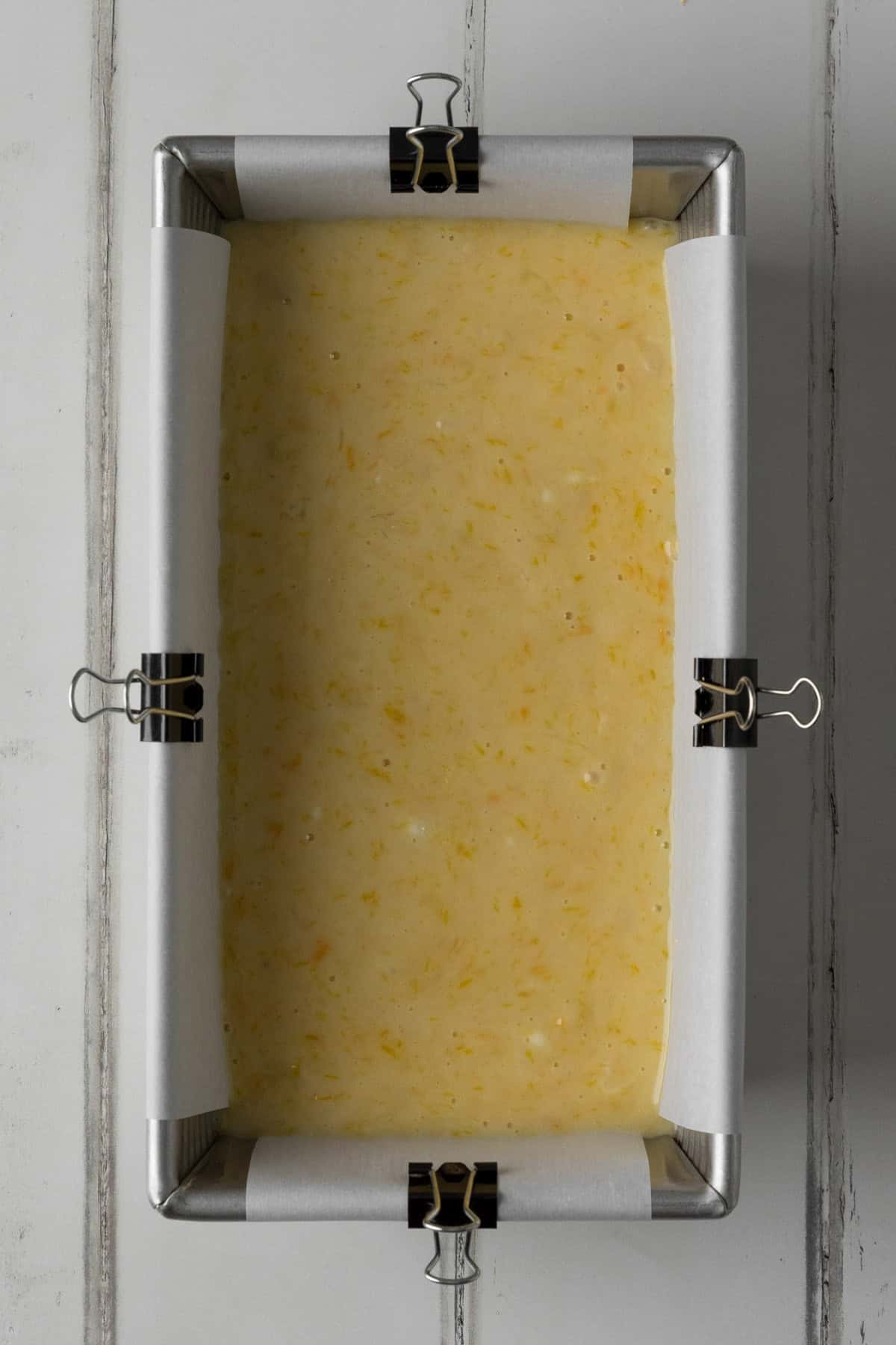 Silver loaf pan lined with white parchment paper, secured with mini binder clips, and filled with orange almond loaf cake batter on a white wood surface.