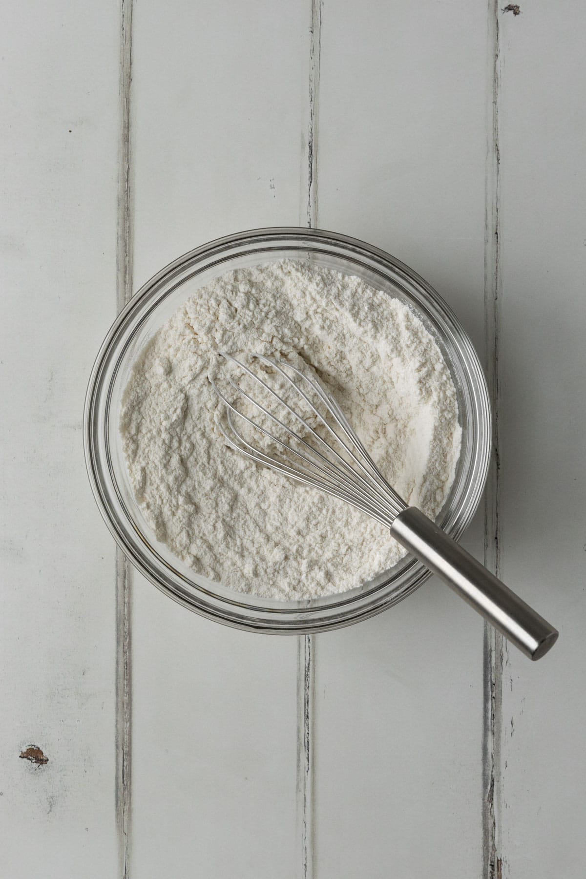 Flour, baking powder, and salt mixed together in a clear bowl with a stainless steel whisk on a white wood surface.