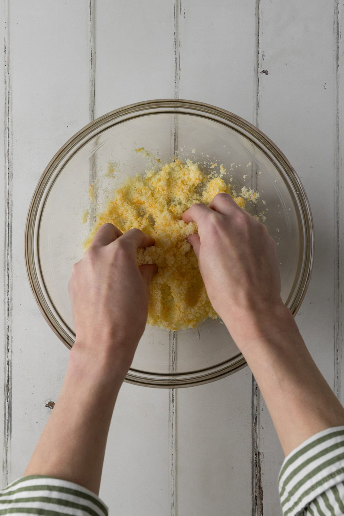 Sugar and orange zest being rubbed together between the fingers of two hands in a clear mixing bowl on a white wood surface.