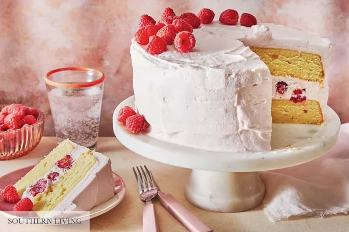 Two-tiered lemon raspberry cake on a white marble cake stand with one slide cut on a pink plate with silverware, a glass of seltzer water, and a bowl of raspberries.